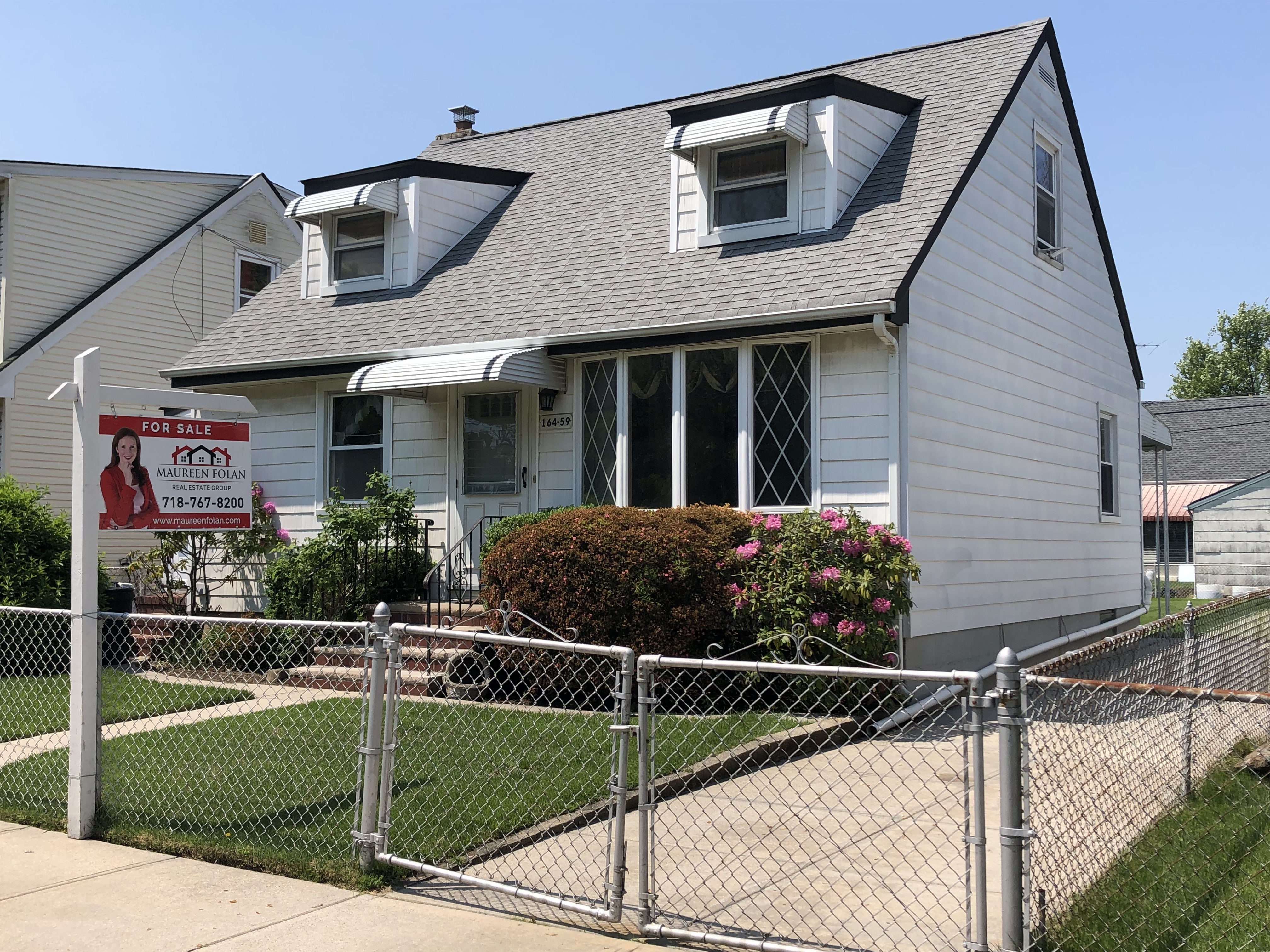 A small white house with a chainlink fence around a neat, square yard has a “For sale” sign in front of it.