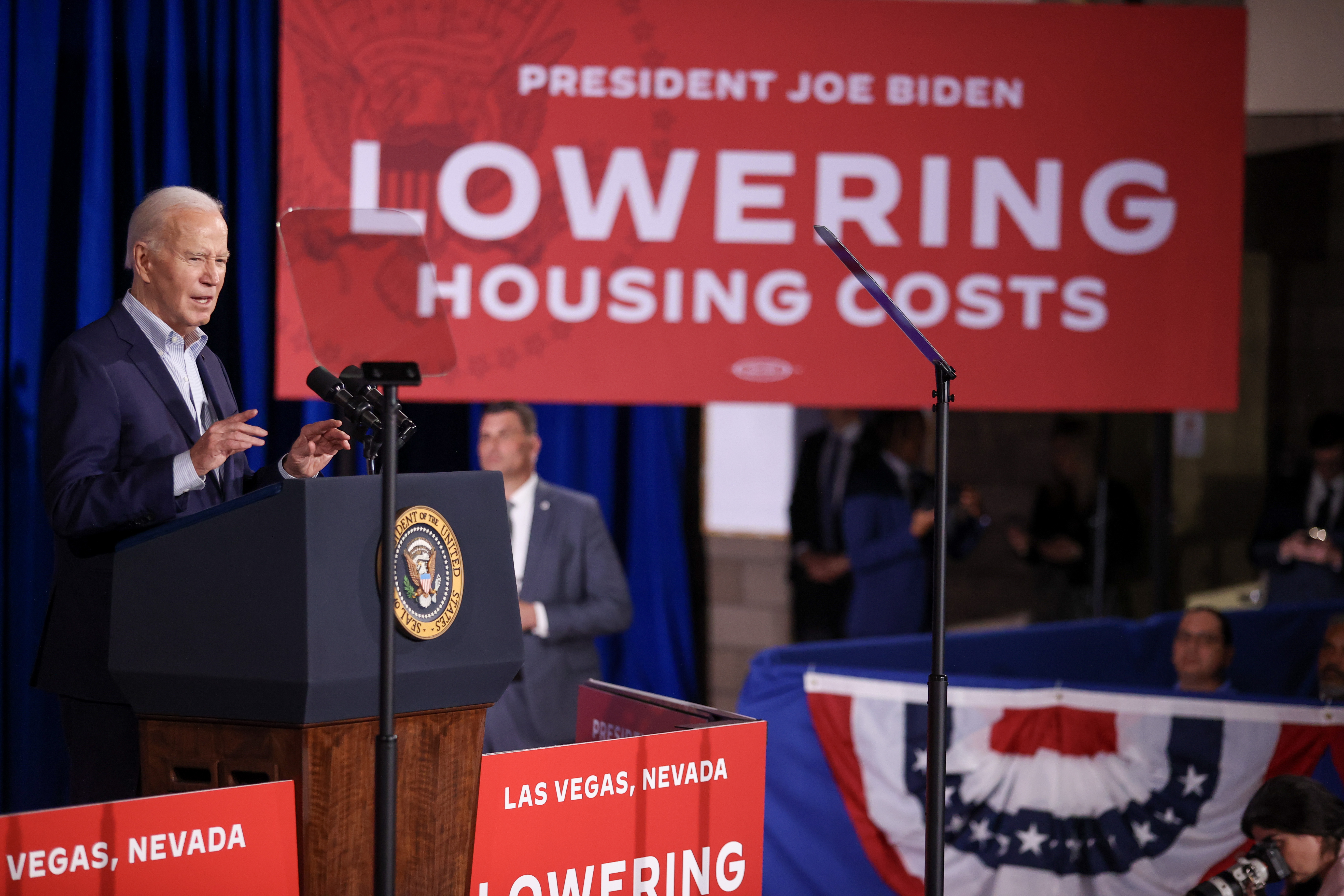 Biden speaks at a podium. Behind him, a large red banner reads: President Joe Biden: Lowering Housing Costs.