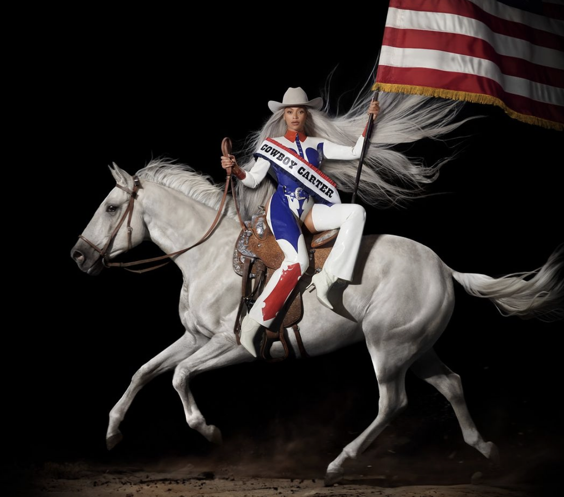 Beyoncé sitting sidesaddle on a galloping white horse, wearing a red, white, and blue Western outfit with a sash reading Cowboy Carter and holding an American flag.