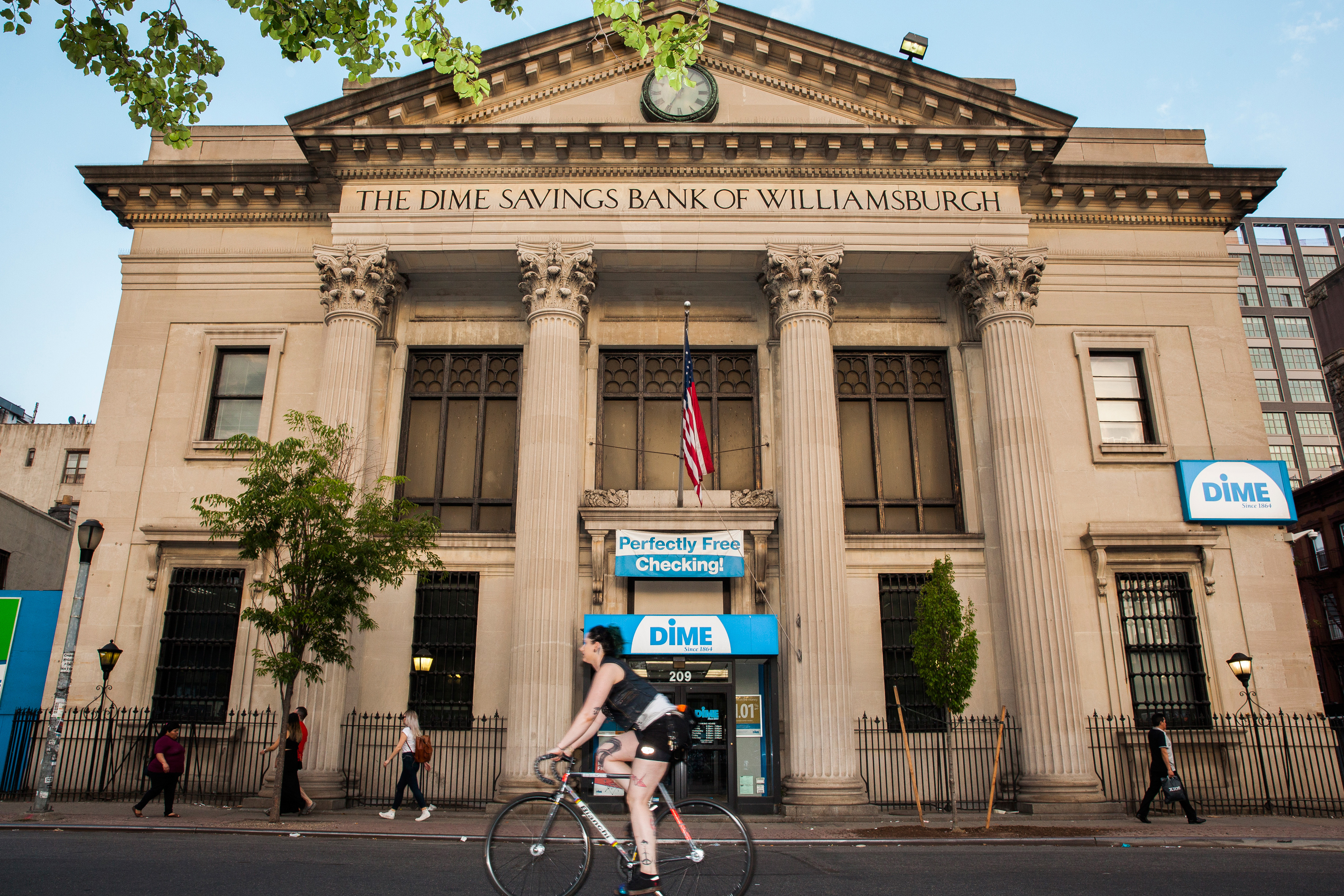 A bicyclist pedals past a stone-fronted bank building, framed by large columns.