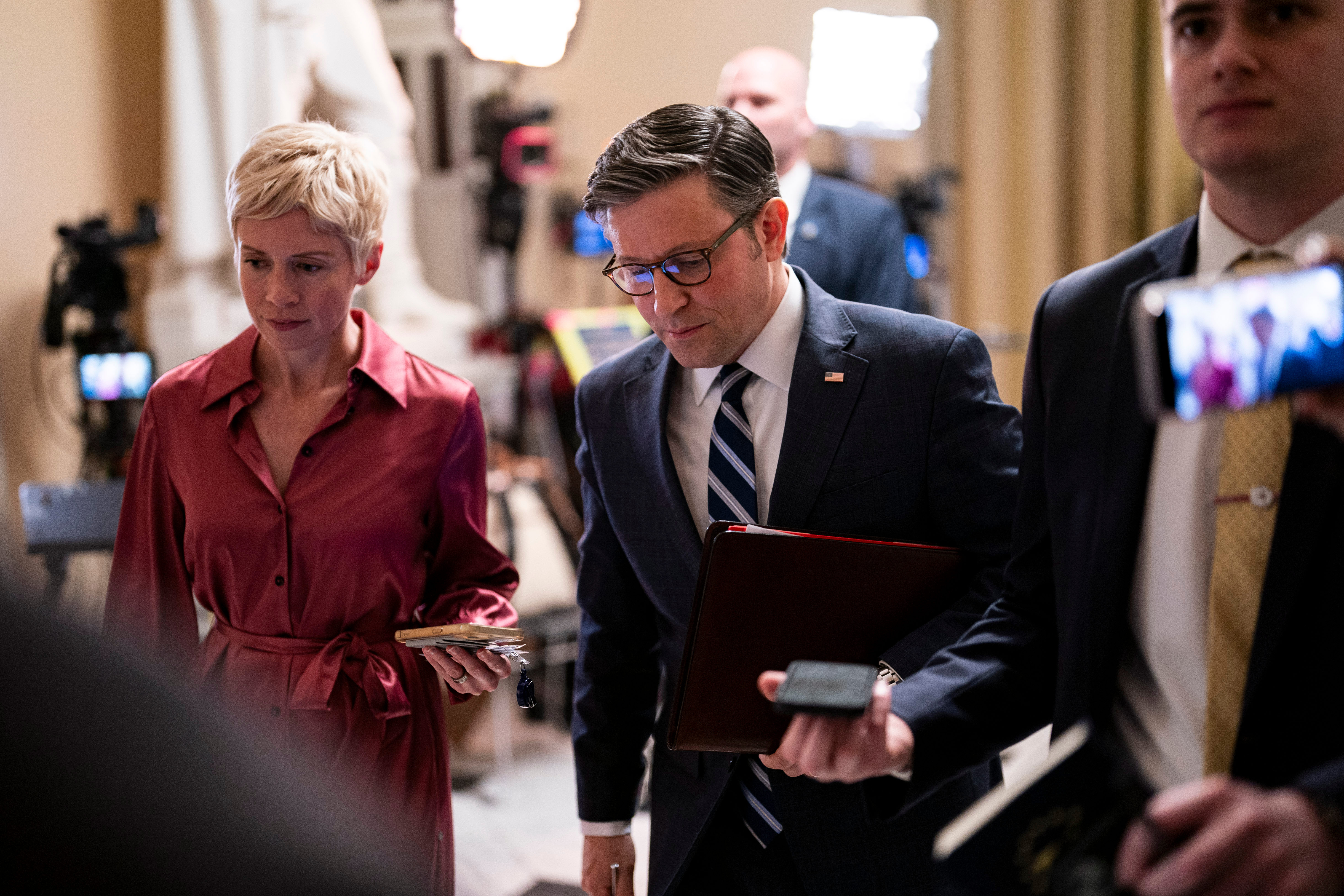 Mike Johnson, in a navy suit and glasses, walks with a leather folder of papers under his arm beside a woman in a burgundy dress.