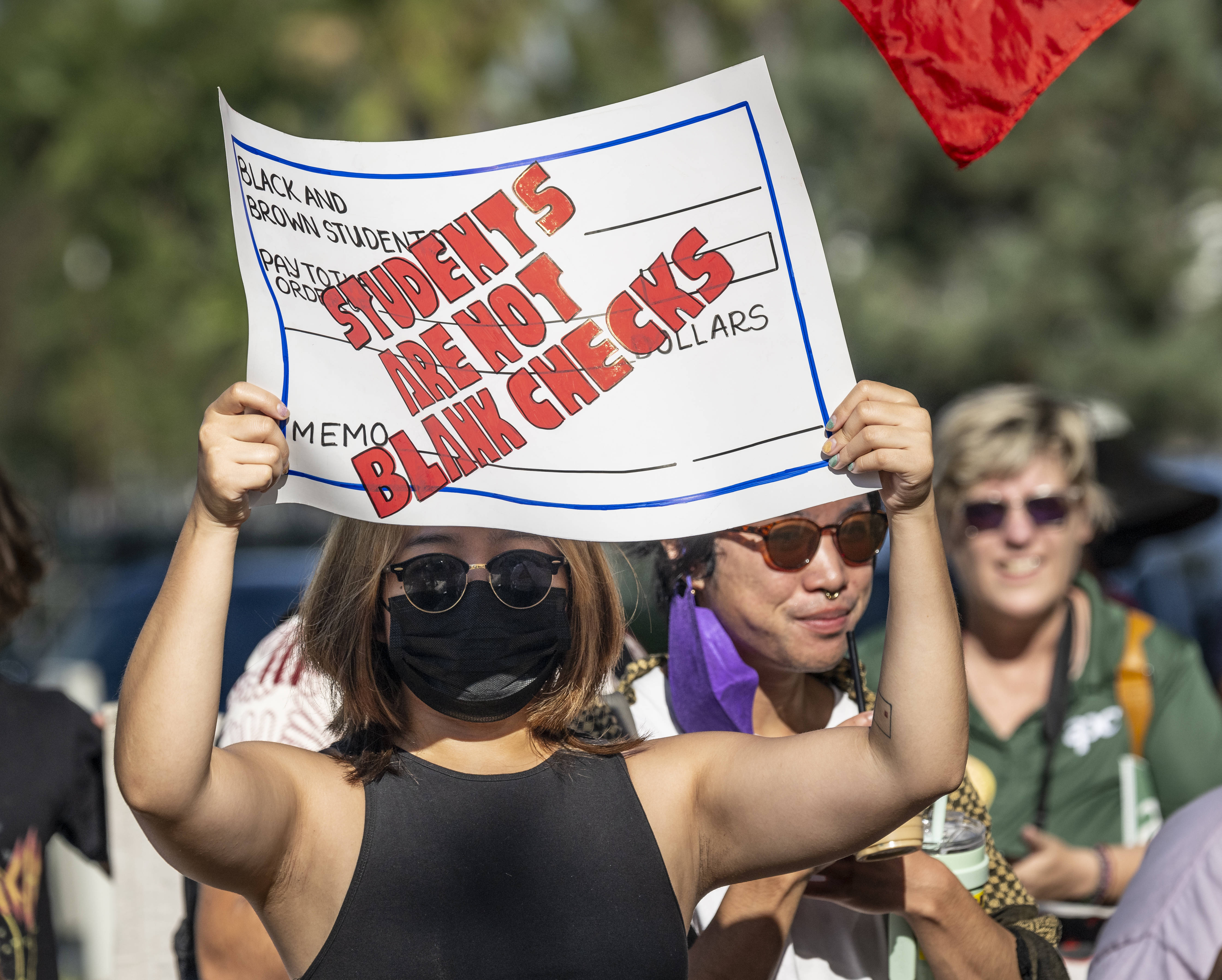 A woman wearing a face-mask and sunglasses holds up a protest sign designed to look like a check that reads “Students are not blank checks” in red letters.