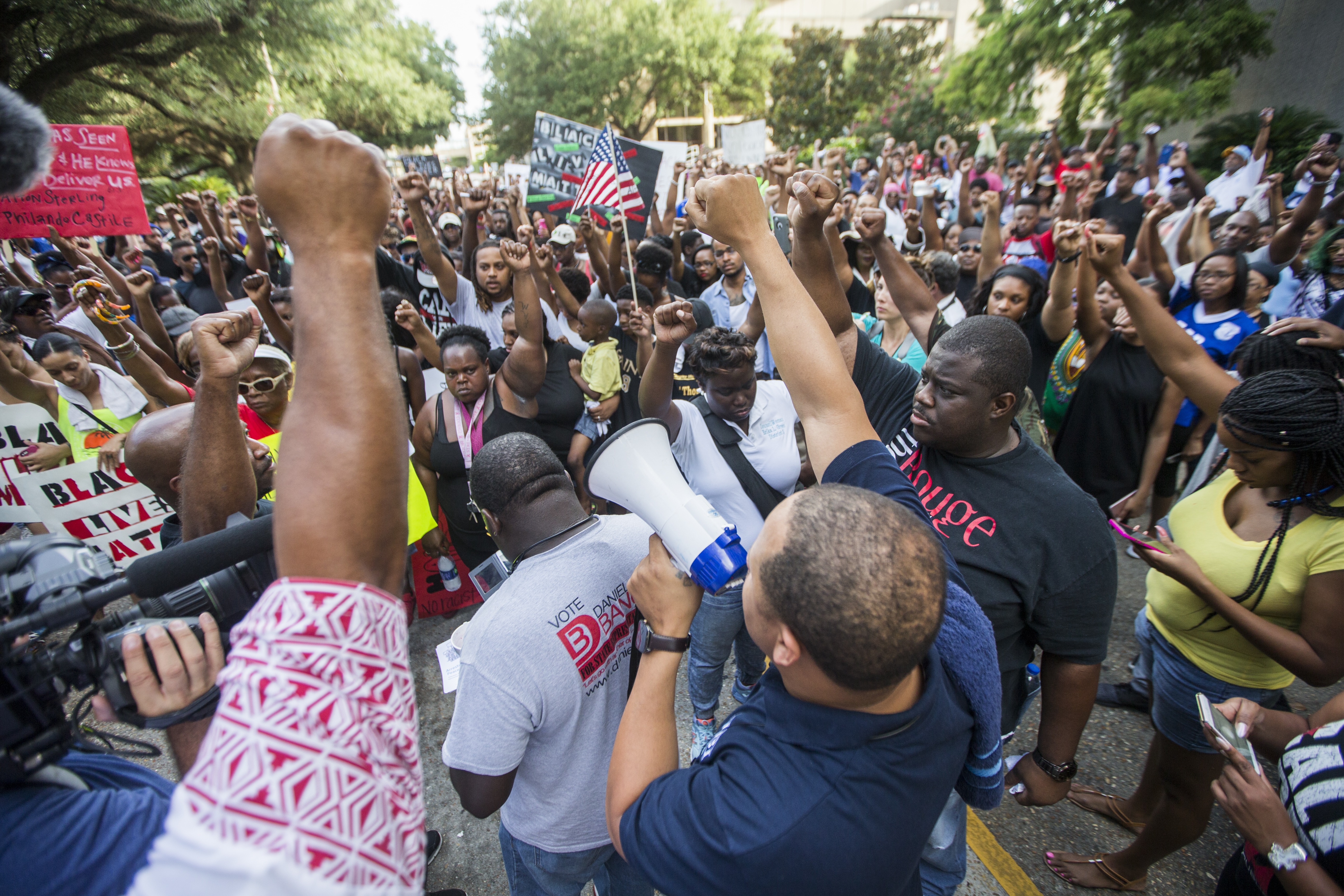 A large crowd of protestors fill the street, many lifting their fists in the air, and one speaking into a megaphone.