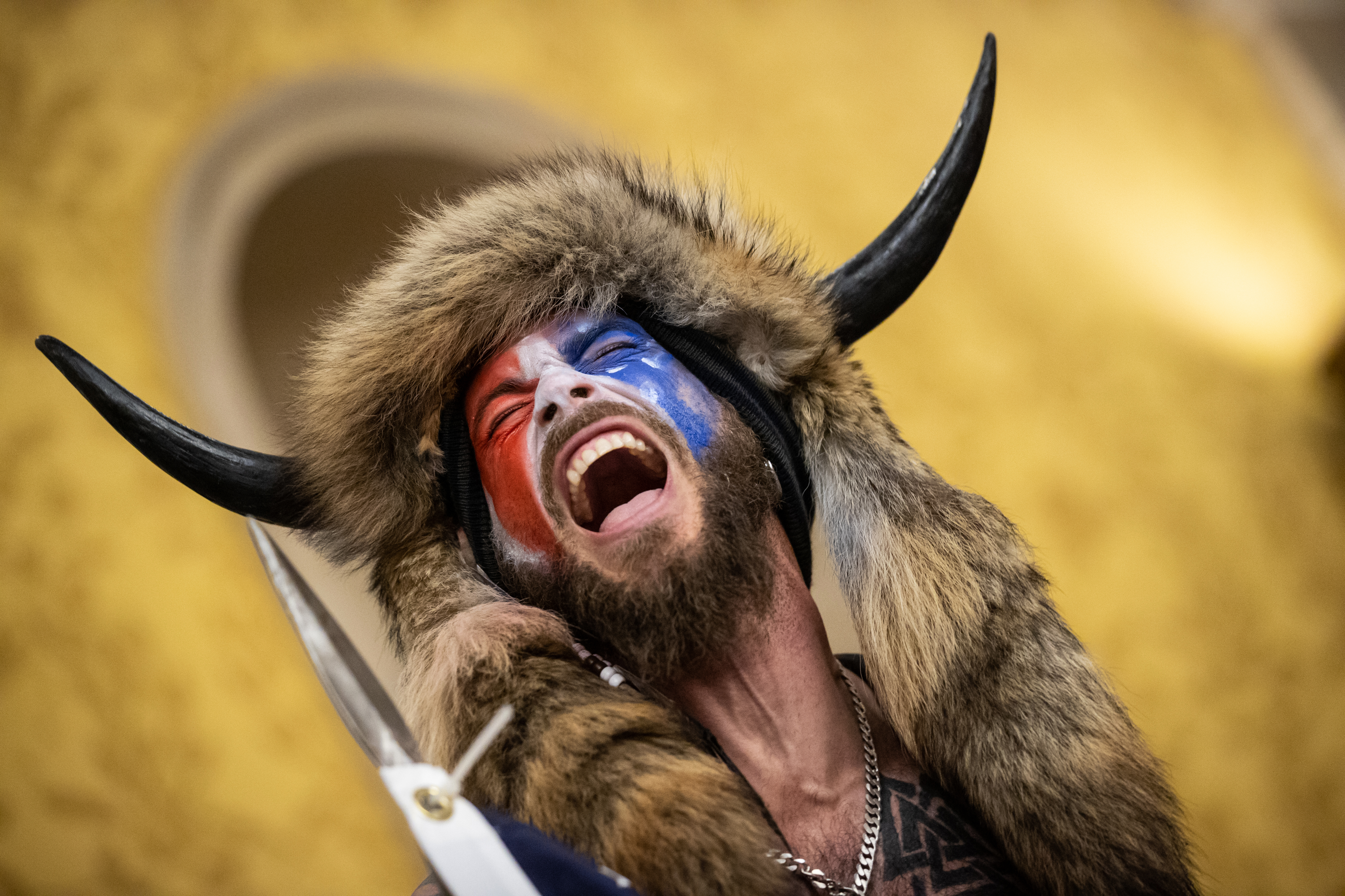A bearded man in red, white, and blue face paint and wearing a furred and horned hat has his mouth open in a scream.