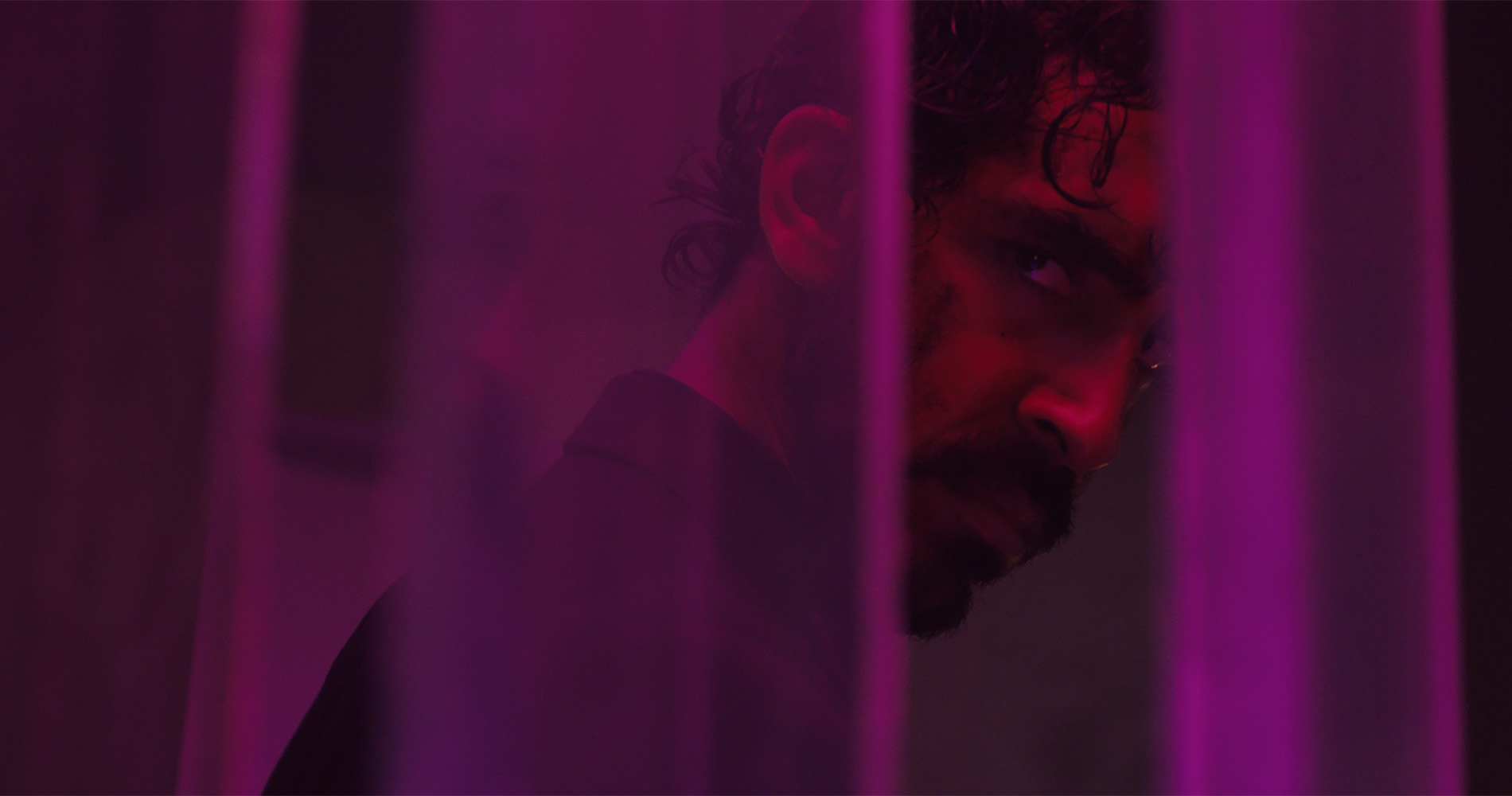 Patel, with a mustache and beard, peers between thin columns of purple glass, his face illuminated by red light.