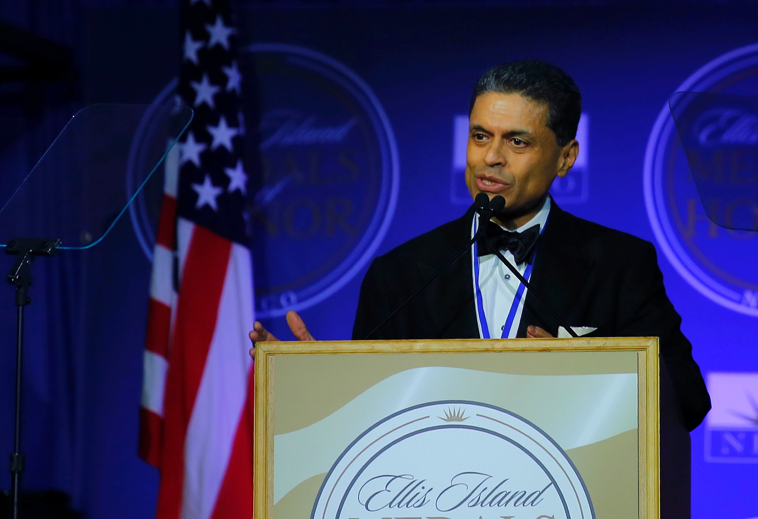 A man wearing a black suit jacket and bowtie, smiling slightly, speaks into a microphone while standing behind a podium. An American flag stands behind him.