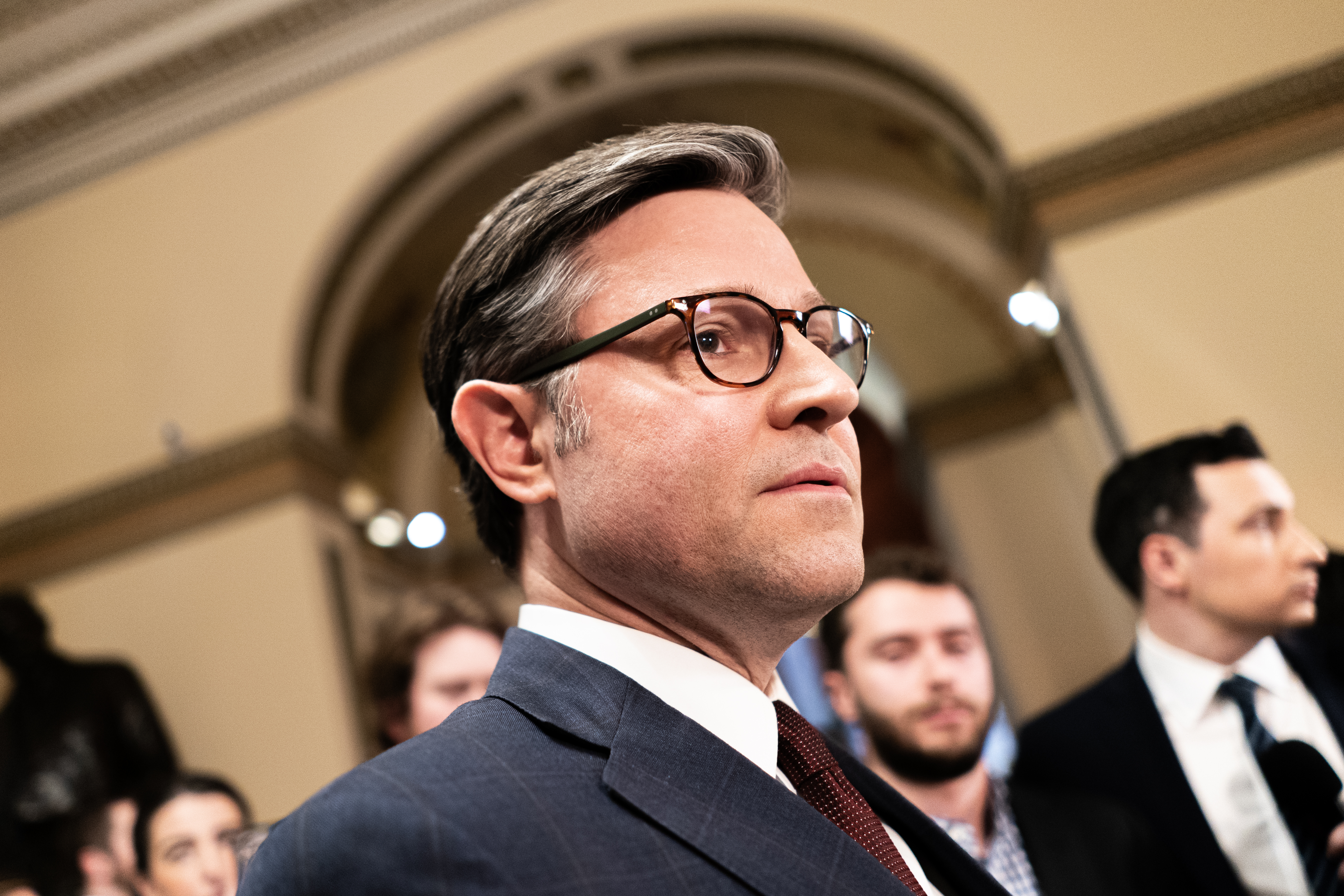 Mike Johnson, in a blue suit and glasses, is seen in profile, looking straight ahead.