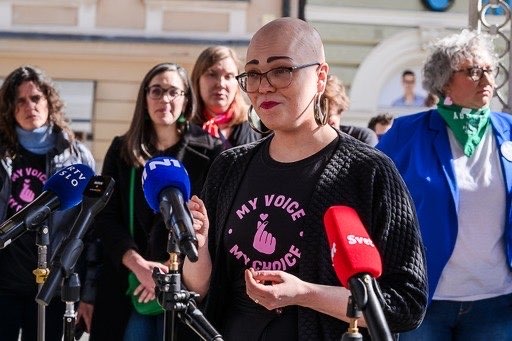 A woman with a shaved head and eyeglasses speaks into several microphones. She wears a shirt with the “My Voice, My Choice” logo. Other people wearing bandana scarves around their necks are gathered behind her.