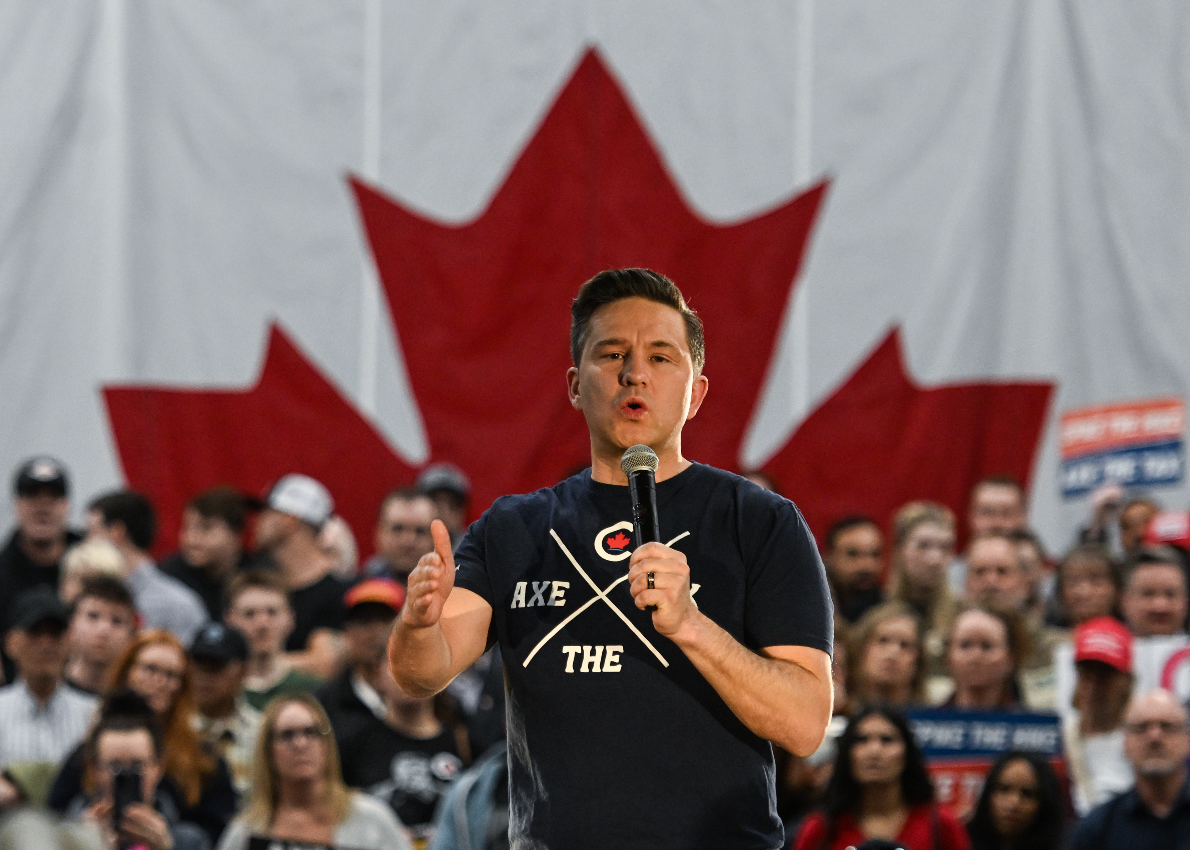 A man in a black T-shirt speaking into a handheld microphone in front of a crowd and a Canadian flag backdrop.