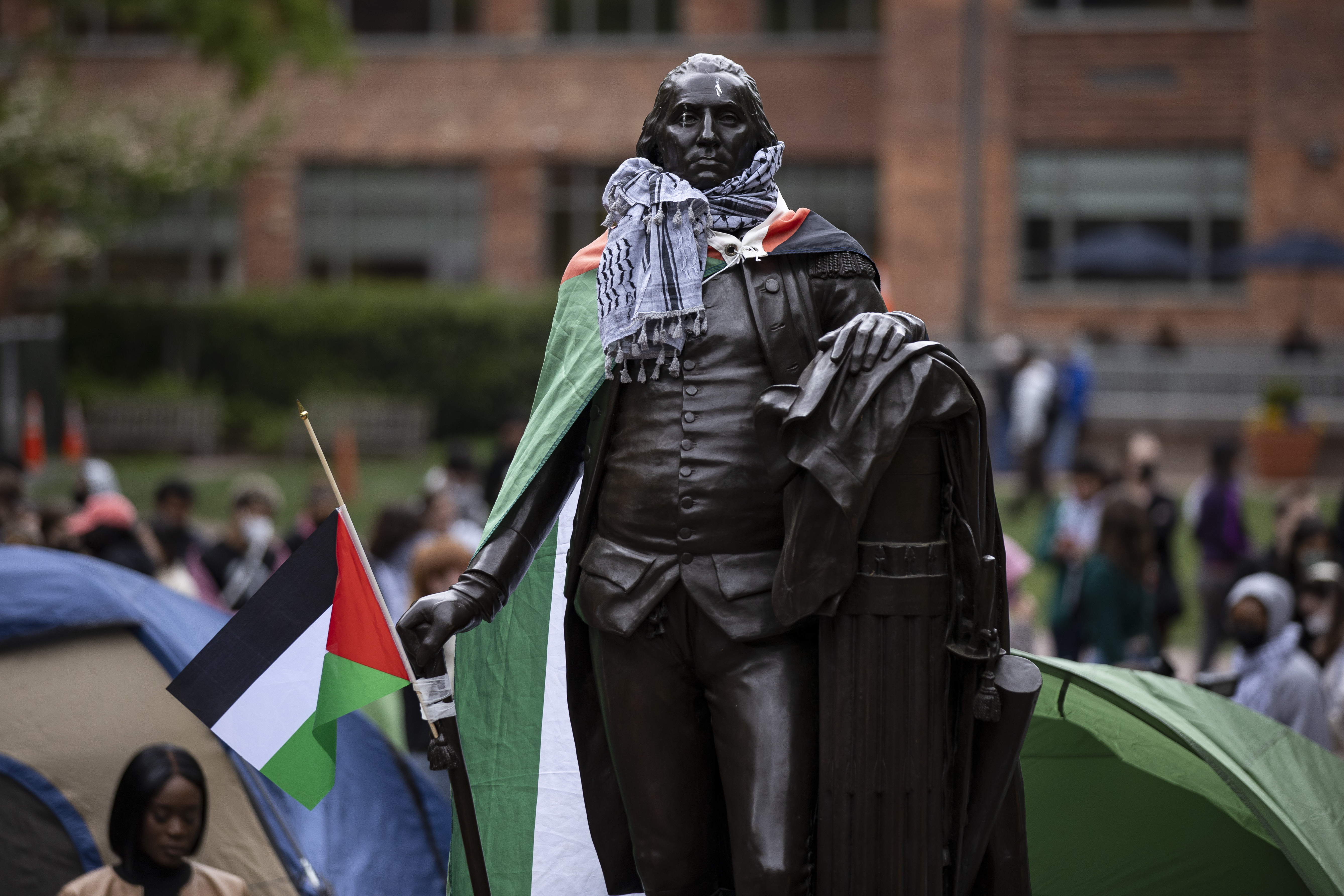 A statue of George Washington has a keffiyeh around its neck and a Palestinian flag as a cape. Behind it, students camp in tents and sit on the grass.