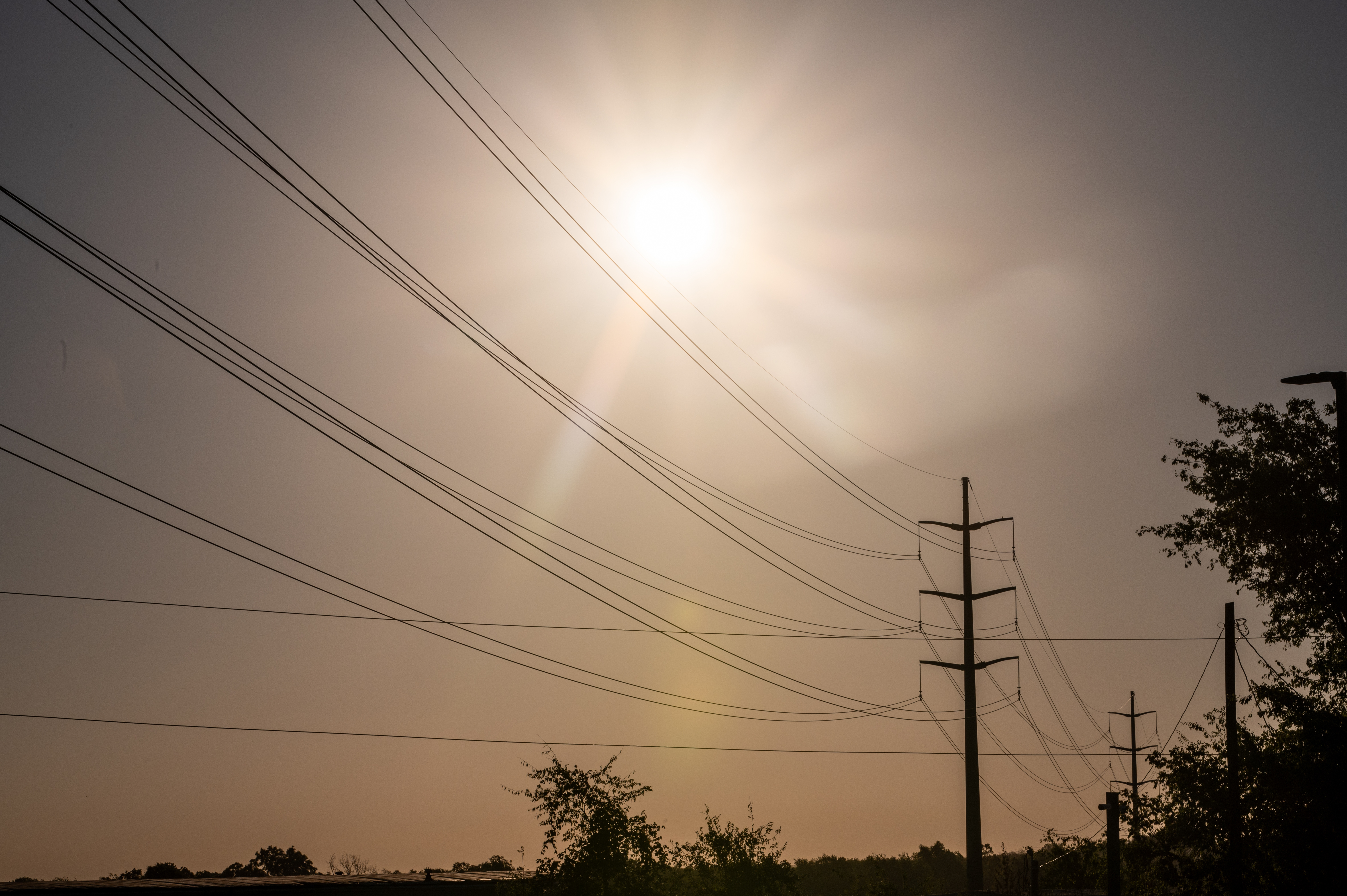 The sun, obscured by a hazy grayish sky, shines above a series of telephone poles and wire.
