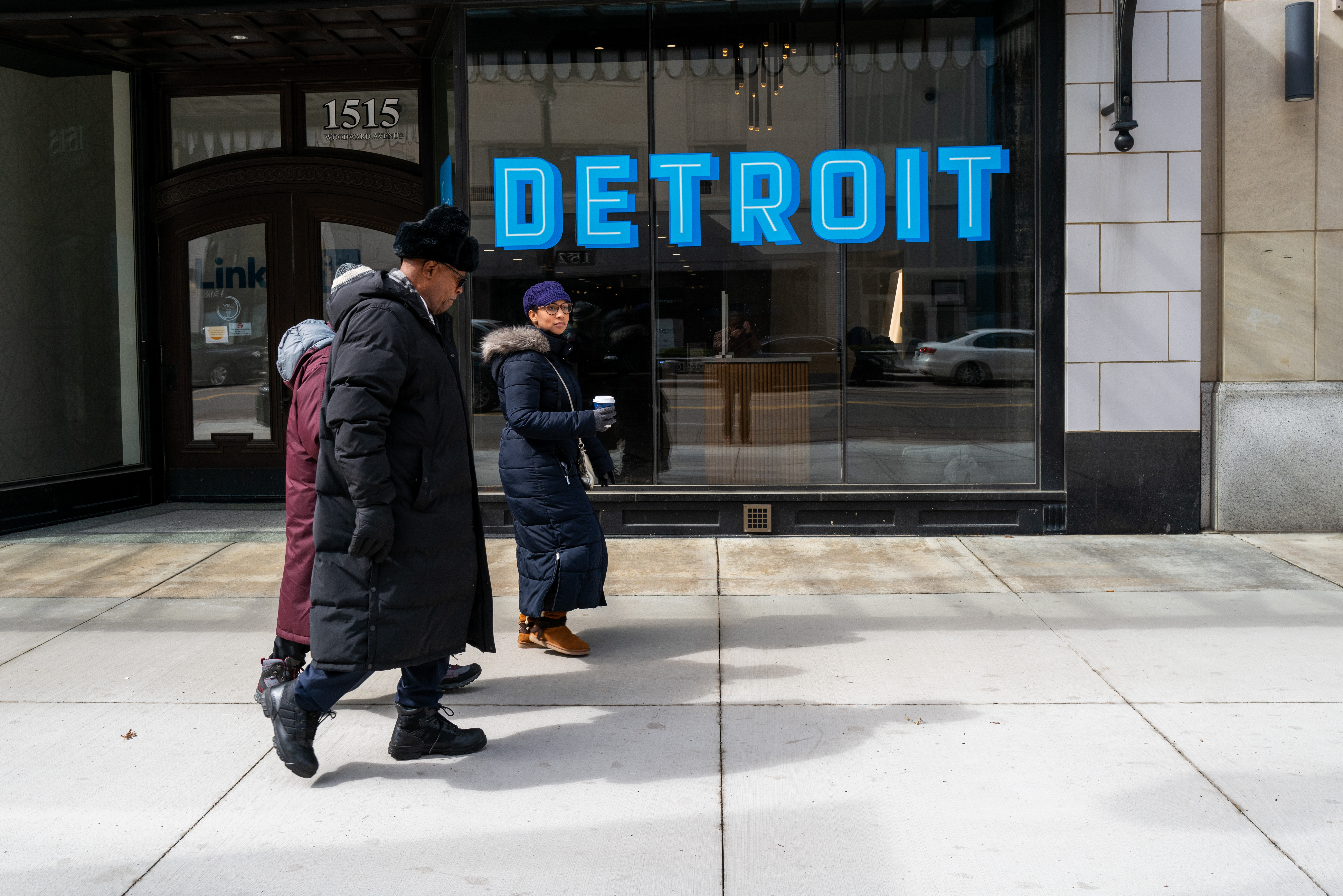 People wearing long puffer winter coats and hats walk on the sidewalk past a building whose window reads DETROIT in big blue letters.