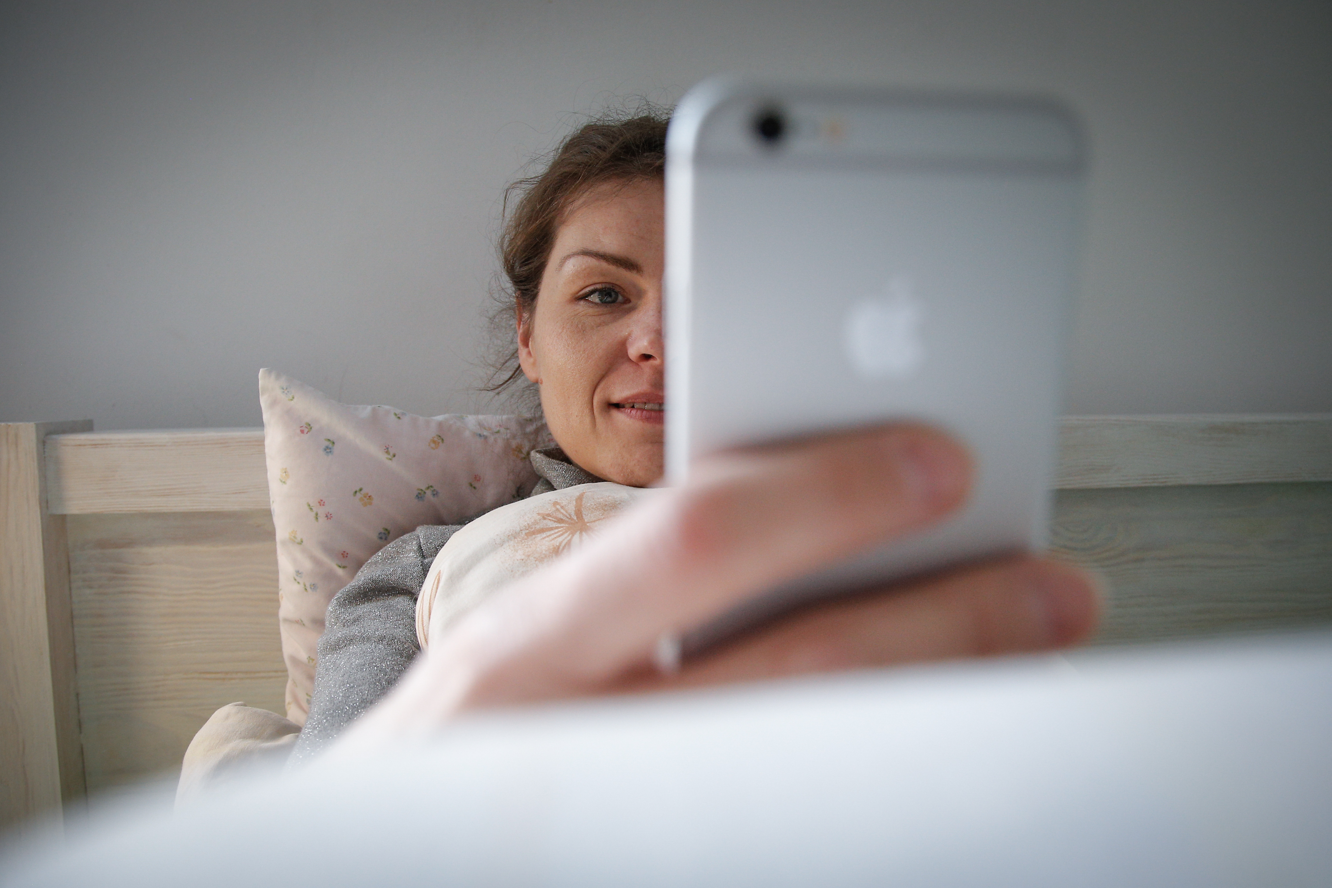 A woman sits in bed, holding her iPhone in front of her face, smiling for the camera.