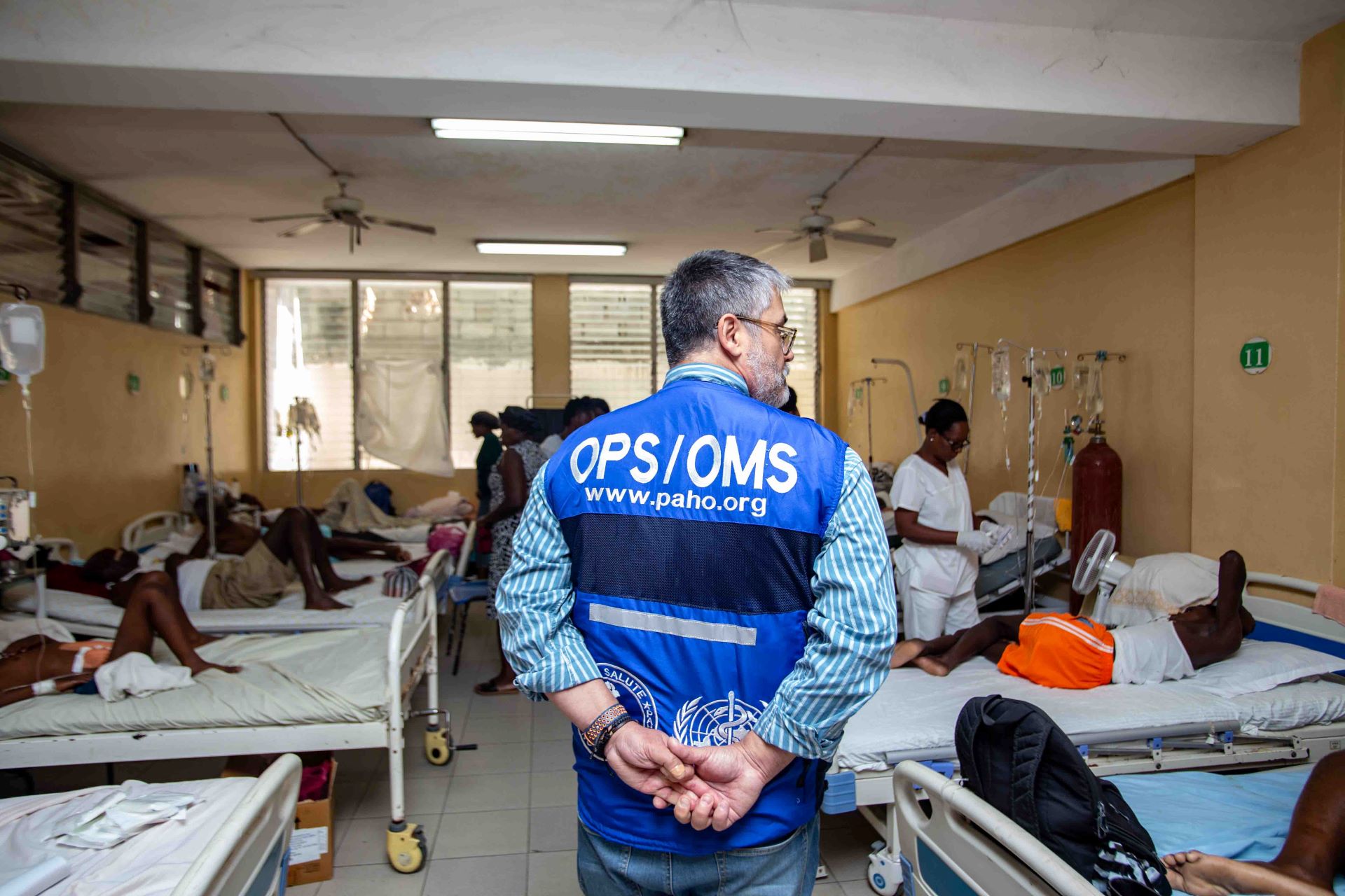 Man in PAHO vest standing in front of beds filled with people in a health centre