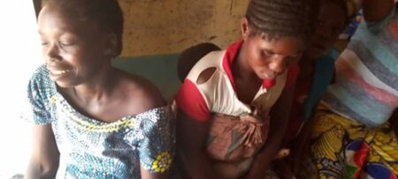Une mère de cinq enfants souriante, vêtue d’une blouse à motifs géométriques, est assise à côté de sa sœur qui porte un enfant sur le dos avec un pagne, dans une pièce modeste.”