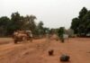 Véhicule des casques bleus marocains de la Minusca traversant une route en terre à la sortie de Bakouma, avec des pierres disposées et un panneau de signalisation, dans un paysage semi-aride.”