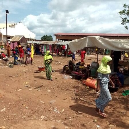 Marché en plein air à Baboua, avec des personnes vendant des marchandises posées à même le sol en terre battue, sous des bâches pour l'ombre, et des vêtements suspendus en arrière-plan"