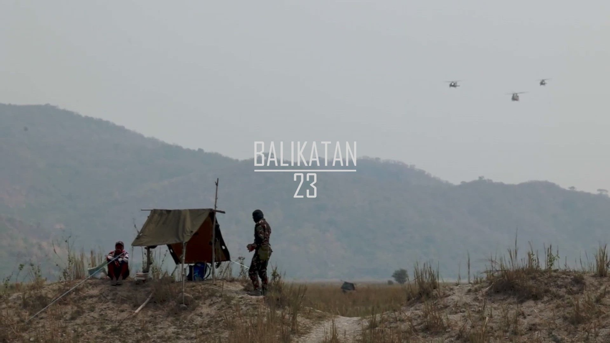 Troops gather by a makeshift structure in a field as helicopters fly overhead. 