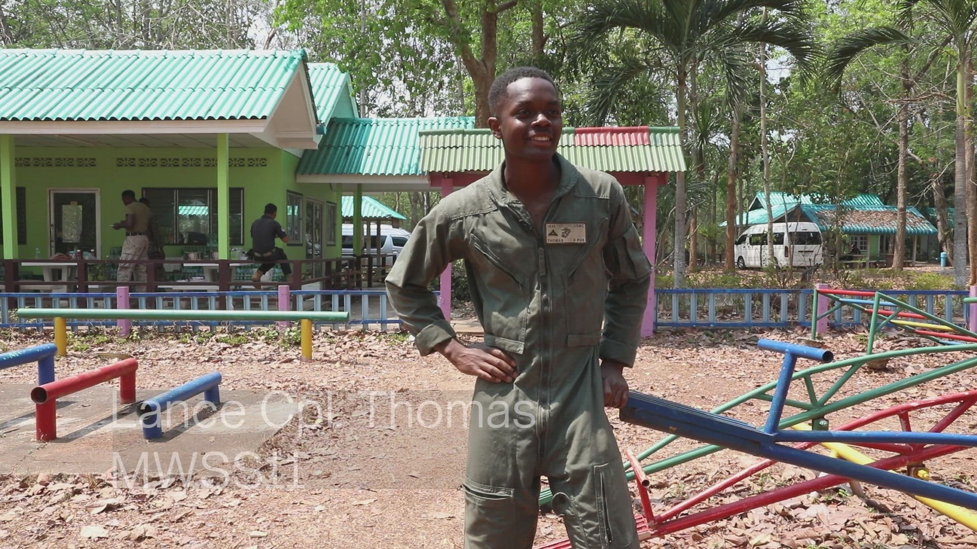 A Marine stands in front of a playground with brightly colored equipment with a clubhouse building in the background.