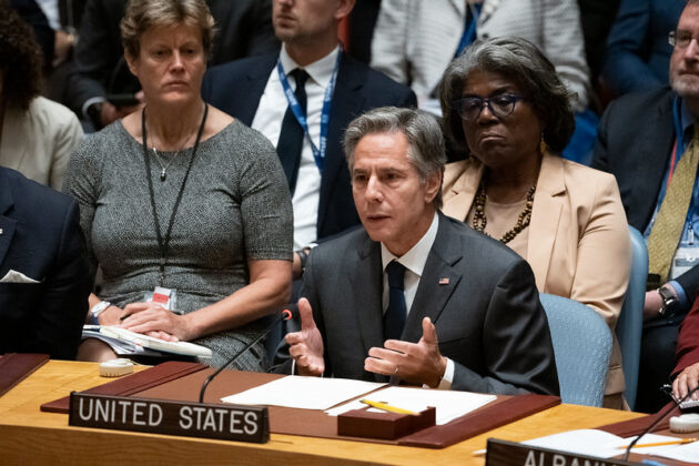 Secretary Blinken sits at a table speaking behind a placard that reads 