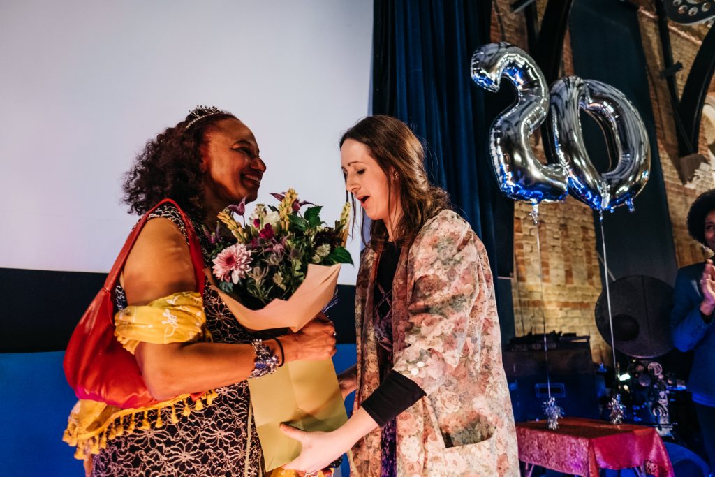 Joanna, a slim white woman with long brown hair hands flowers to Artistic Director Maria Oshodi as they stand smiling in front of a large balloon reading 20