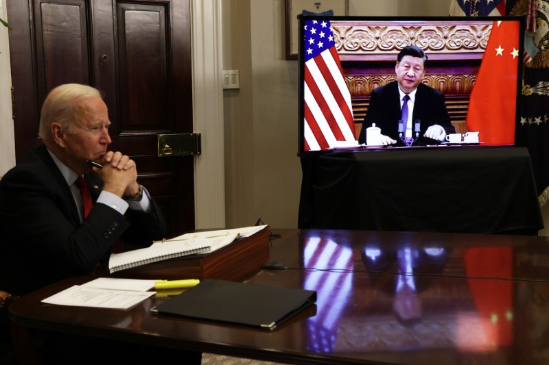 U.S. President Joe Biden sits at a table next to a screen showing with Chinese President Xi Jinping during a virtual meeting at the White House.