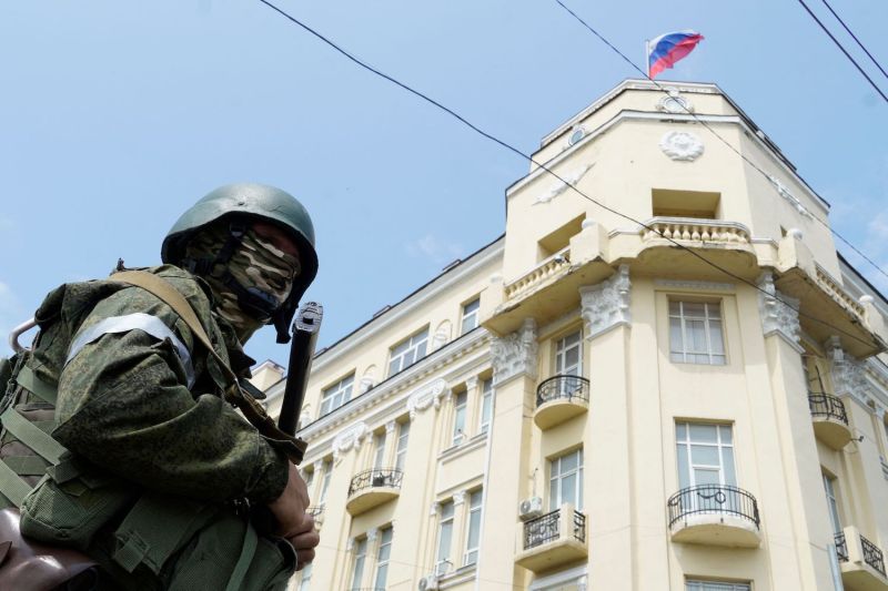 A member of the Wagner Group stands guard with a rifle in front of a large stucco building flying a Russian flag against a blue sky. The guard wears a green helmet and uniform along with a camouflage face covering.