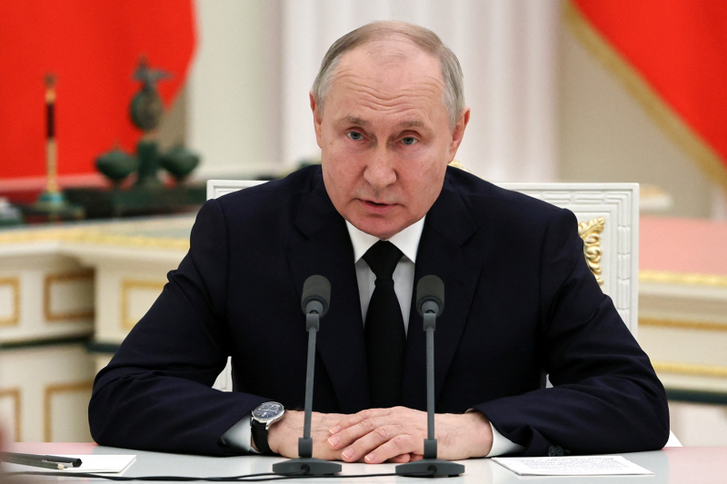 Russian President Vladimir Putin, a 70-year-old man wearing a black suit and tie, leans forward with his hands on the table in front of him as he speaks into a microphone.