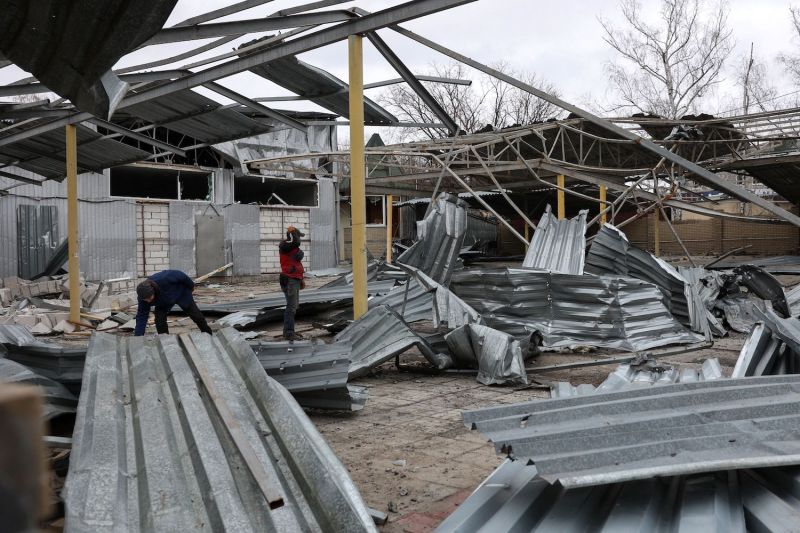 Two workers stand among the rubble of a damaged bus station. Crumpled sheets of corrugated metal surround them on the ground, and the windows of the station building behind them have been shattered and blown out.