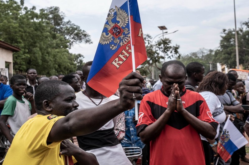 A crowd of residents of Bangui, Central African Republic, demonstrate on the street on a cloudy day. In the foreground, a man in a yellow shirt waves a Russian flag. Next to him, another man bows his head with his hands clasped, possibly in prayer.