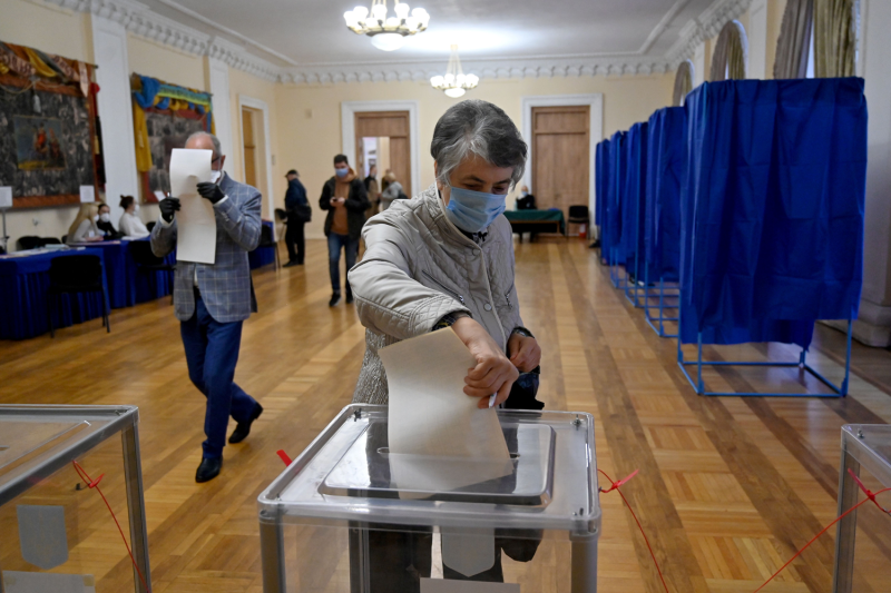 A middle-aged woman wearing a surgical face mask and holding a purse inserts a paper into a transparent ballot box. Behind her is a room with cream walls and sculpted crown moldings, and other people move around between a table of election staffers and curtained voting booths.