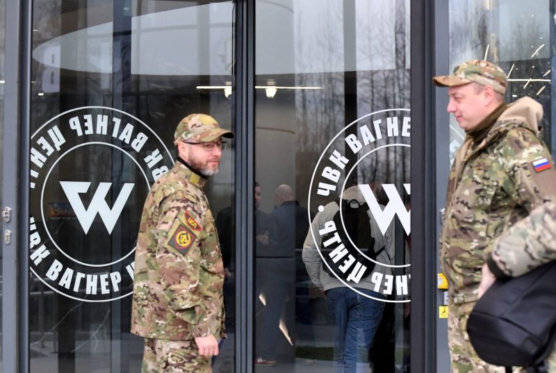 Two men wearing military camouflage fatigues and matching hats stand in front of a building with large glass doors labeled with a logo that reads "Wagner Center" in Russian.