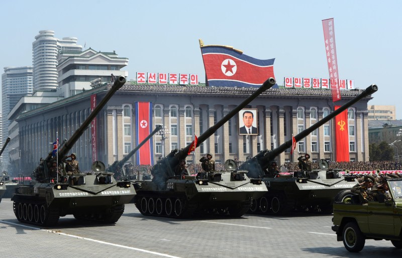 Three tanks lined side-by-side in a row parade down a broad paved boulevard. Soldiers in green uniforms and helmets stand on the tanks and salute. Behind them is a large building fronted with columns, North Korean flags, and a portrait of Kim Il Sung.