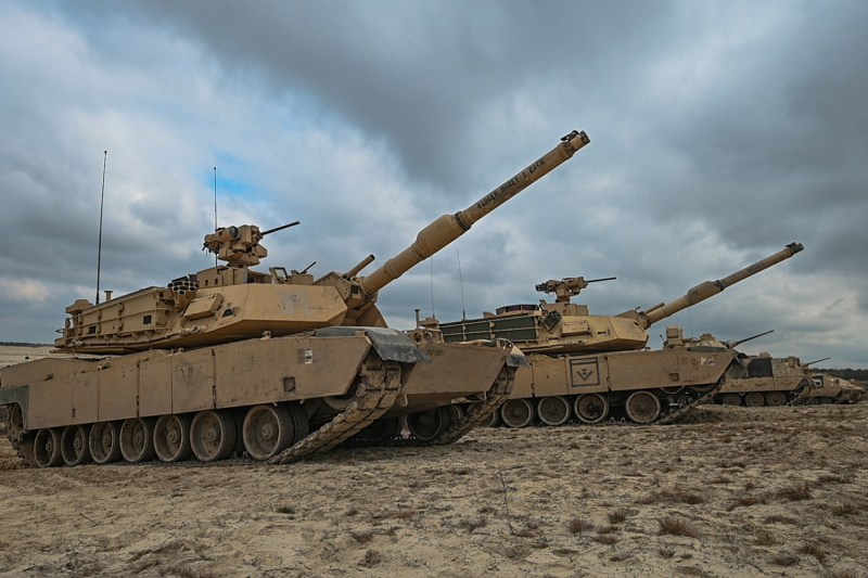 Two camel-colored Abrams tanks move across a sandy landscape beneath a cloudy sky.