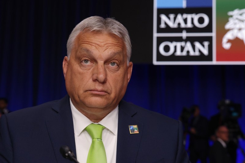 Hungarian Prime Minister Viktor Orban glances up to something off-camera, frowning slightly. Orban is a 60-year-old man with white hair, wearing a dark suit and lime green tie. Other attendees stand behind him, in front of a curtain and a sign bearing the NATO logo.