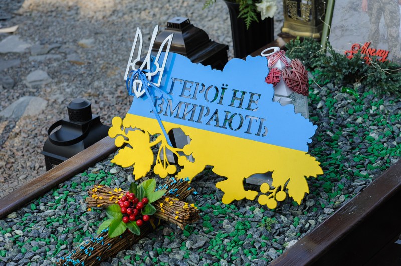 A sign in the shape of the map of Ukraine in the blue and yellow colors of the flag with the inscription "Heroes do not die" in Cyrillic rests on the grave of a fallen soldier at Lychakiv cemetery in Lviv, Ukraine. A cross made of sticks and greenery and lanterns frame the scene.