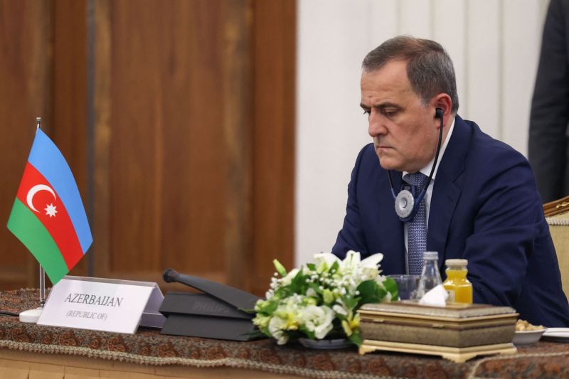 Azerbaijani Foreign Minister Jeyhun Bayramov sits at a desk and looks down at its surface during a diplomatic meeting. Bayramov wears a dark blue suit, and a microphone sits on his desk between a small Azerbaijani flag and a bouquet of white flowers.