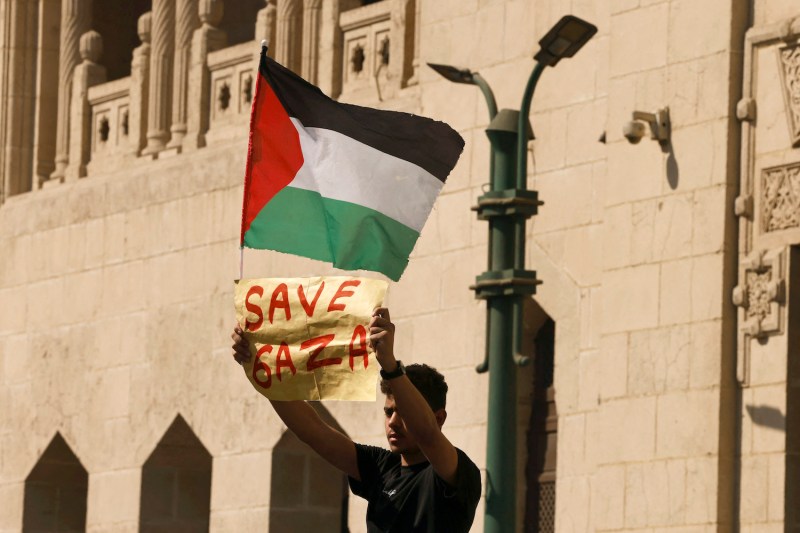 A young man holds up a sign that reads “Save Gaza” in handwritten red paint. He also holds a Palestinian flag as he stands in front of the stone walls of a mosque in Cairo.