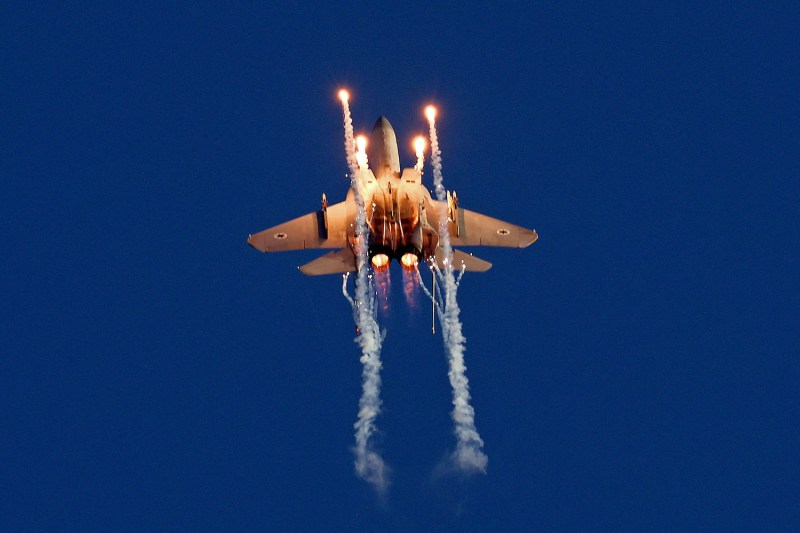 Flares and smoke contrails extend from an Israeli F-15 Eagle fighter in flight against a blue sky.