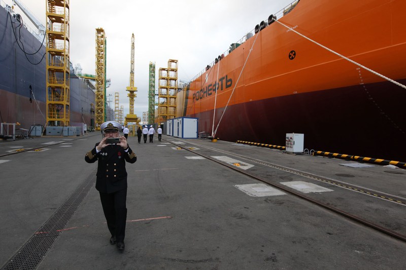 A captain in full uniform and hat uses a cell phone to take a photo during a ceremony to name a Rosneft oil tanker in Russia. A large ship and scaffolding extends behind him as people walk in the distance at the port.