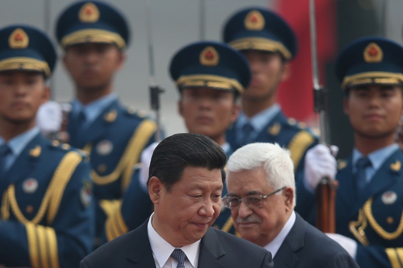 Chinese President Xi Jinping walks closely in front of Palestinian Authority President Mahmoud Abbas in front of a group of Chinese soldiers wearing formal uniforms. Both officials wear dark suits and look down at something out of view.