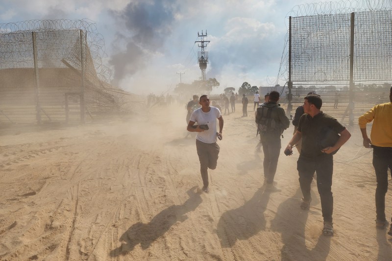 Palestinians run across a dusty landscape through a breach in a high wire fence topped with barbed wire on the Israel-Gaza border.