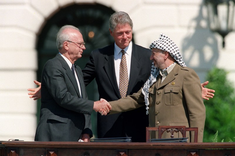 U.S. President Bill Clinton, in a dark suit and tie, smiles as he stands between Palestine Liberation Organization leader Yasser Arafat, in a uniform and checked headscarf, and Israeli Prime Minister Yitzahk Rabin, in a dark suit, as they shake hands in front of the White House. Clinton's arms are outstretched as he gestures around the other two men.