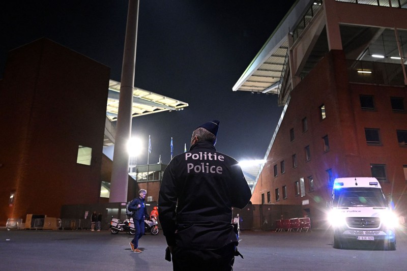 A police officer wearing a uniform and hat is seen from behind as he stands guard outside the King Baudouin Stadium in Brussels. A police vehicle with headlights on is in front of him and lights and the roof of the soccer stadium are seen in the distance.