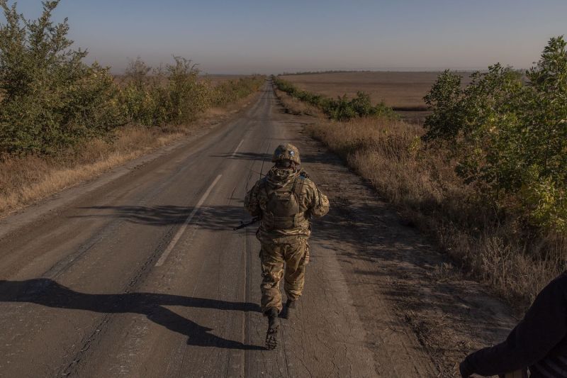 A Ukrainian soldier wearing a camouflage military uniform is seen from behind as they walk down a cracked paved road surrounded by grassy fields and open sky. The soldier is holding a rifle and casts a long shadow.