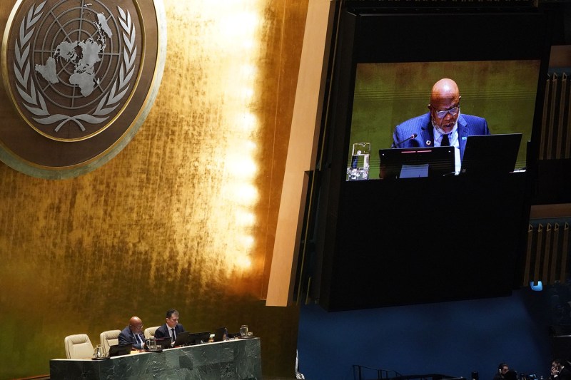 U.N. General Assembly President Dennis Francis reads aloud from a laptop screen while sitting at a podium desk in the U.N. meeting chamber. Francis, a middle-aged man wearing a blue suit, is also visible on a video screen hung on the wall next to a large United Nations logo.