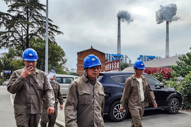 Four workers in blue hardhats walk past a coal plant. One is smoking a cigarette.