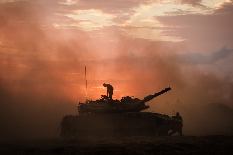 A soldier stands atop an Israeli battle tank near the border of Gaza. They are silhouetted against a an orange smoky sky.