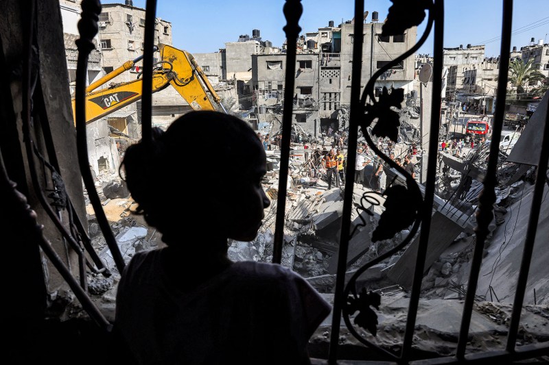 A young girl is silhouetted as she stands behind the metal mesh that covered the window of a building that was hit by Israeli bombardment in the southern Gaza. In front of her is a scene of destruction with the rubble of crumbled buildings and an excavator sifting through the piles.