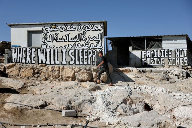 A young man walks across a ground of cracked stone in front of a cluster of small buildings below a clear blue sky. The buildings are covered with text painted in Arabic and English. One sign reads "Families, not firing zones," and another reads, "Where will I sleep?"